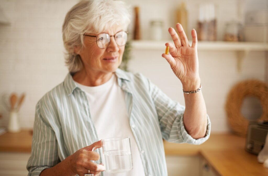 An older adult woman with glasses and white hair holding a vitamin e tablet in her left hand a glass of water in her right hand