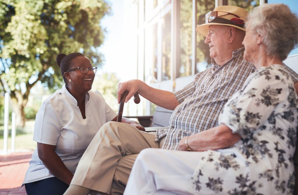 A senior couple sits on a bench under the sun while speaking cheerfully to a nurse.