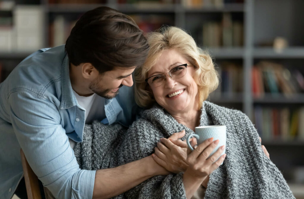 A young man embracing his elderly mother while she sits sipping coffee from a cup in her hand.