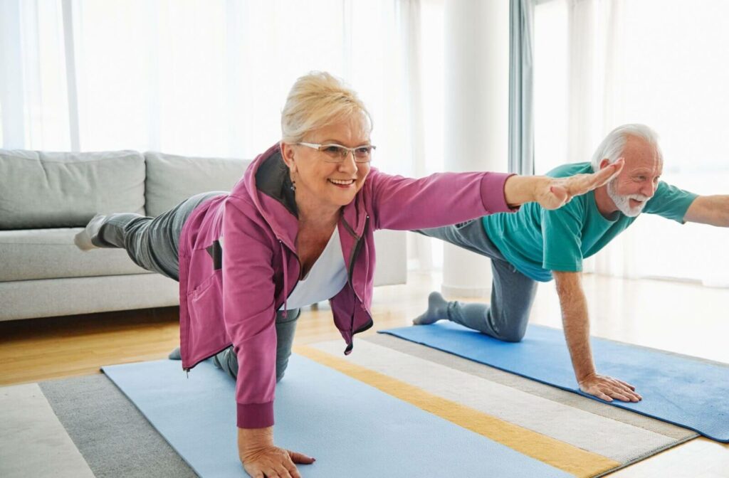 Two seniors stretching in a well-lit room with blue curtains, standing with arms extended overhead in a side stretch