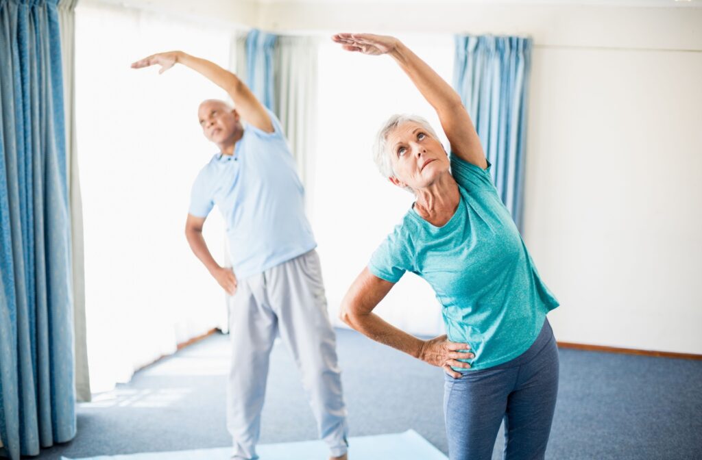 Two older adults performing a balance exercise on yoga mats in a bright living room, extending one arm and one leg