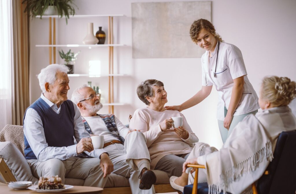 Several seniors have an afternoon coffee in a communal area of their assisted living community while a nurse pats a resident on the shoulder.