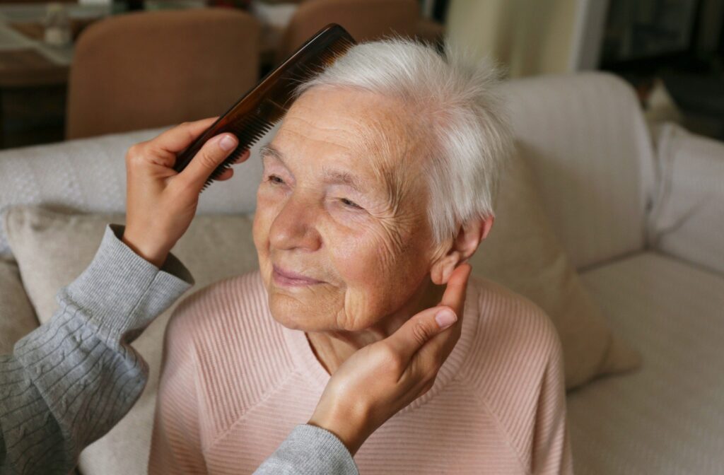 An older adult smiles while sitting on their bed as an out-of-frame caregiver carefully combs their hair